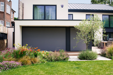 Load image into Gallery viewer, A motorized external roller blind in a dark color, fully extended and installed on a house facade, with a garden in the foreground.
