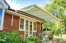 Load image into Gallery viewer, A beige patio awning installed over a brick house's entrance, with trees and part of a garden visible behind it.